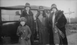 [Three women and a girl in hats and wintercoats standing in front of a car]